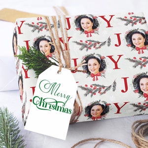 May include: White gift wrap with a repeating pattern of red "J" letters, green holly sprigs, red berries, and a white snowflake design. The gift wrap features a photo of a person in each of the "J"s. The gift wrap is tied with twine and has a white gift tag with the words "Merry Christmas" in green cursive.