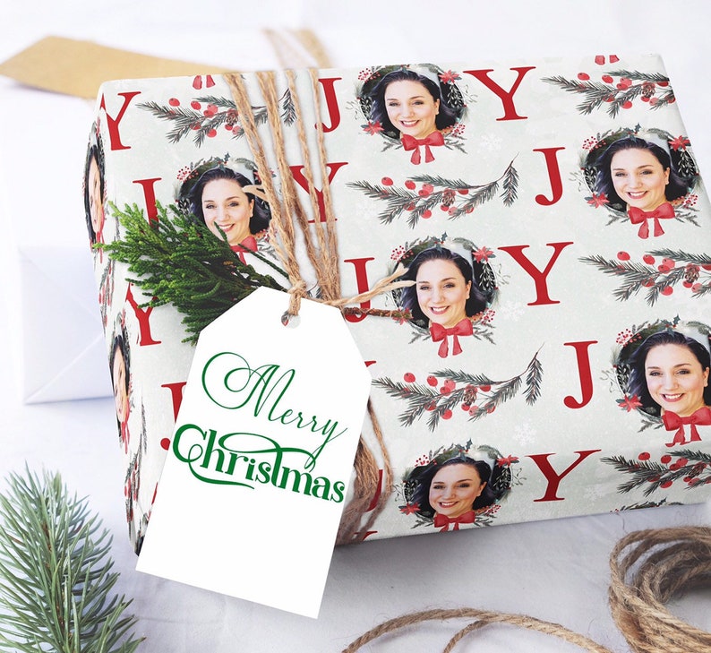 May include: White gift wrap with a repeating pattern of red "J" letters, green holly sprigs, red berries, and a white snowflake design. The gift wrap features a photo of a person in each of the "J"s. The gift wrap is tied with twine and has a white gift tag with the words "Merry Christmas" in green cursive.