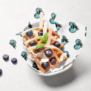Pu&ograve; includere: Tre waffle guarnite con mirtilli e zucchero a velo, servite in una ciotola bianca con un rivestimento di carta a motivi blu e bianchi. Il rivestimento di carta ha un motivo ripetuto di cuori con il testo "Sweet Impact" all'interno di ogni cuore.