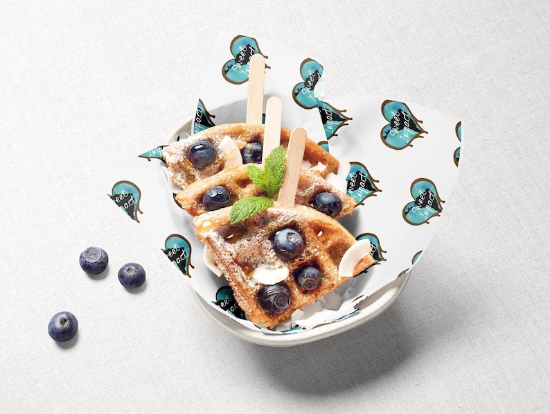 Pu&ograve; includere: Tre waffle guarnite con mirtilli e zucchero a velo, servite in una ciotola bianca con un rivestimento di carta a motivi blu e bianchi. Il rivestimento di carta ha un motivo ripetuto di cuori con il testo "Sweet Impact" all'interno di ogni cuore.