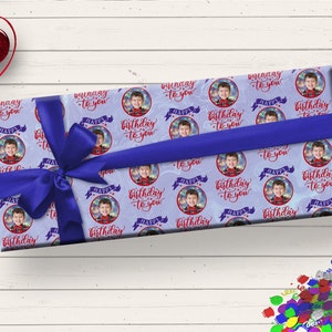 May include: A blue gift box with a blue ribbon wrapped around it. The box is covered in a wrapping paper with a repeating pattern of a boy's face in a circle with the text "Happy Birthday to you" in red.