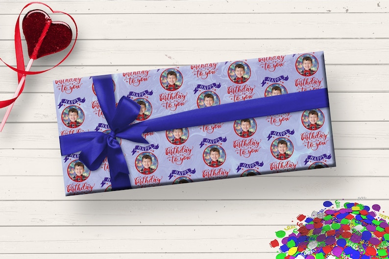 May include: A blue gift box with a blue ribbon wrapped around it. The box is covered in a wrapping paper with a repeating pattern of a boy's face in a circle with the text "Happy Birthday to you" in red.