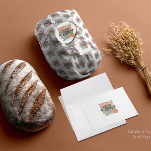 May include: A loaf of dark crusty bread, a package wrapped in white paper with a repeating Johnny Cakes Bakery logo and peach graphic, a white envelope, a white note card with the same logo, and a bundle of dried wheat stalks.