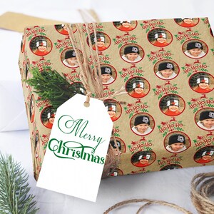 May include: A brown gift wrap with a repeating pattern of red circles with green holly and the text "Ho-Ho-Ho". Each circle contains a photo of a child wearing a hat. A white gift tag with green text that reads "Merry Christmas" is attached to the gift wrap.