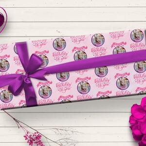 May include: Pink gift wrap with a repeating pattern of a girl's photo in a circle with the text "Happy Birthday to you" in pink lettering. The gift is wrapped with a purple ribbon.