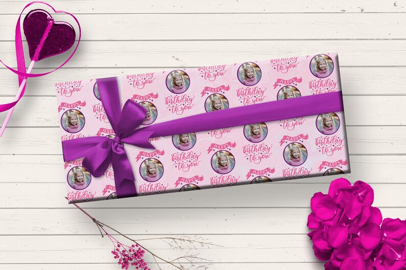 May include: Pink gift wrap with a repeating pattern of a girl's photo in a circle with the text "Happy Birthday to you" in pink lettering. The gift is wrapped with a purple ribbon.
