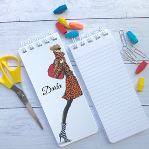 May include: Two spiral bound notebooks. One notebook has a white background with a black and white illustration of a woman wearing a leopard print coat and carrying a red bag. The notebook has the name "Darla" written on it. The other notebook has a white background with lined pages.