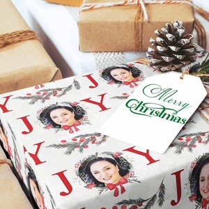 May include: White gift wrap with a repeating pattern of red "J" letters, green pine branches, red berries, and a photo of a woman. The gift wrap has a white gift tag with green text that reads "Merry Christmas".