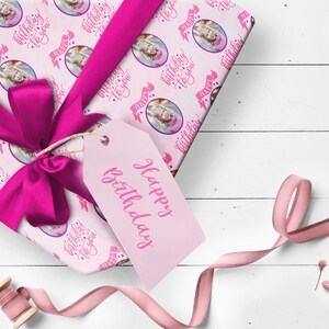 May include: Pink gift wrap with a repeating pattern of a girl's face in a circle with the text "Happy Birthday to you" in pink script. The gift is wrapped with a pink ribbon and a pink gift tag that says "Happy Birthday".