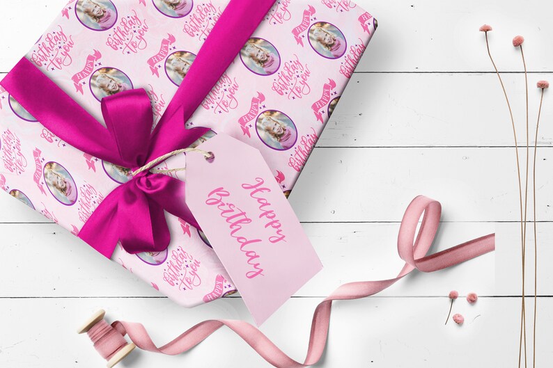 May include: Pink gift wrap with a repeating pattern of a girl's face in a circle with the text "Happy Birthday to you" in pink script. The gift is wrapped with a pink ribbon and a pink gift tag that says "Happy Birthday".
