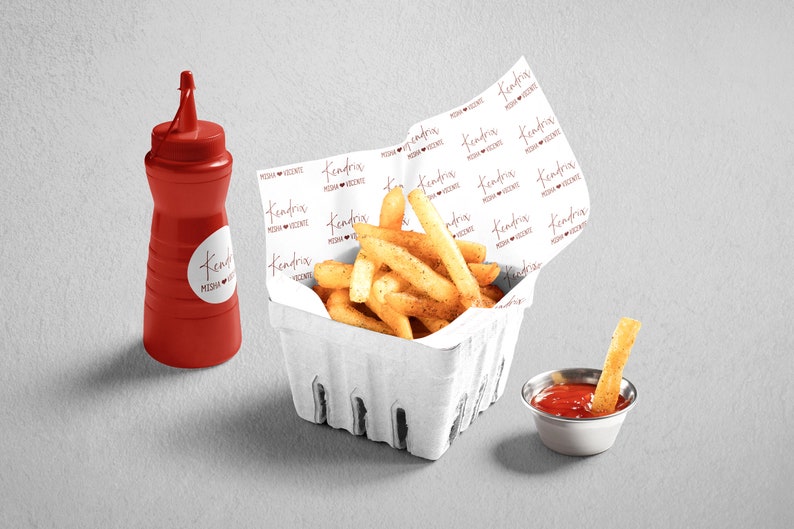 Pu&ograve; includere: Un cestino di carta bianco pieno di patatine fritte dorate. Il cestino &egrave; rivestito di carta bianca stampata con le parole "Kendra's Pizza & Vicente" in un font corsivo rosso. Una piccola bottiglia di ketchup di plastica rossa e una piccola ciotola di metallo con ketchup sono sul tavolo.