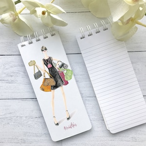 May include: A white spiral bound notepad with a colorful illustration of a woman carrying multiple handbags on the front cover. The back cover is blank with lined paper. The notepad has the name "Heather" written on the front cover.