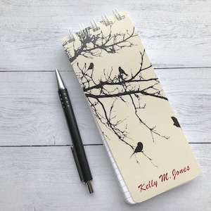 May include: A spiral bound notebook with a cream background and black silhouette of branches and birds. The notebook has the name "Kelly M. Jones" written in red on the bottom right corner.