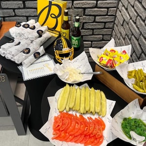 May include: A catering spread featuring wrapped sandwiches, a yellow and black football helmet, two beer bottles, and bowls containing sliced pickles, tomato slices, chopped onions, yellow peppers, and green relish, all on branded paper.