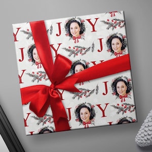May include: White wrapping paper with a red ribbon and a repeating pattern of the word "JOY" in red letters, pine branches, red berries, and a woman's face in a wreath.