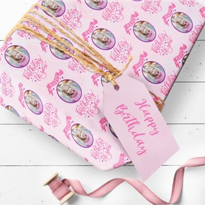 May include: Pink wrapping paper with a repeating pattern of a girl's photo in a circle with the text "Happy Birthday to you" in pink script. The paper is wrapped around a gift box with a pink gift tag that says "Happy Birthday".