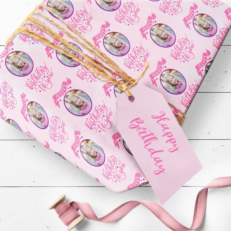 May include: Pink wrapping paper with a repeating pattern of a girl's photo in a circle with the text "Happy Birthday to you" in pink script. The paper is wrapped around a gift box with a pink gift tag that says "Happy Birthday".