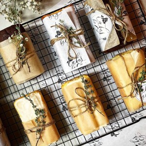 Puede incluir: Primer plano de una rejilla de alambre con varios panes de molde envueltos en papel marr&oacute;n y atados con cuerda. Los pasteles est&aacute;n decorados con ramitas de tomillo y tienen etiquetas escritas a mano que dicen "Cakes by ...".