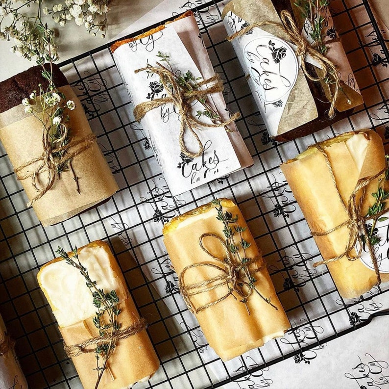 Puede incluir: Primer plano de una rejilla de alambre con varios panes de molde envueltos en papel marr&oacute;n y atados con cuerda. Los pasteles est&aacute;n decorados con ramitas de tomillo y tienen etiquetas escritas a mano que dicen "Cakes by ...".