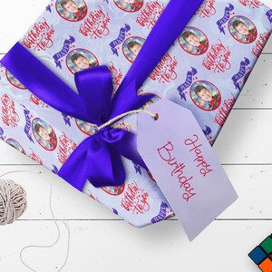 May include: A gift wrapped in blue and white wrapping paper with a blue ribbon. The wrapping paper has a repeating pattern of a child's face in a circle with the text "Birthday to you" in red. The gift is tied with a blue ribbon and has a white tag with the text "Happy Birthday" in red.