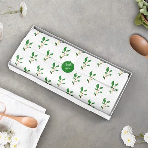 Puede incluir: Una caja de regalo blanca con un patr&oacute;n floral y una pegatina verde que dice "Gracias". La caja est&aacute; sobre una superficie gris.