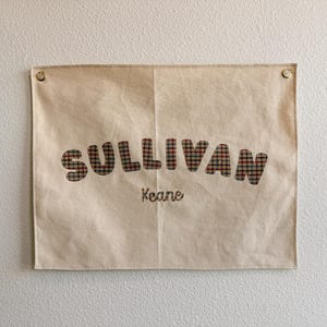 May include: A beige fabric wall hanging with the name "SULLIVAN" in plaid letters. Below the name, the word "Keane" is embroidered in a smaller font. The hanging is secured to the wall with two gold-colored tacks.