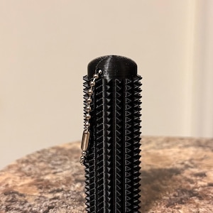 May include: A black, cylindrical object with a textured, spiky surface and a small chain with a clasp. The object is standing upright on a textured, brown surface. The object appears to be 3D printed.