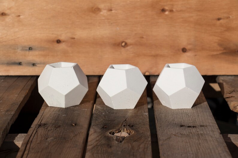May include: Three white geometric concrete planters with a faceted design. The planters are arranged in a row on a wooden surface.