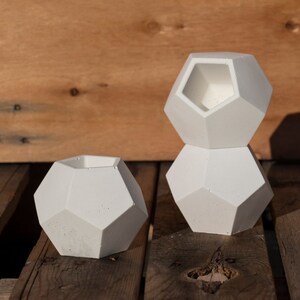 May include: Three white geometric concrete planters of different sizes. The planters are stacked on top of each other. The planters are sitting on a wooden surface.