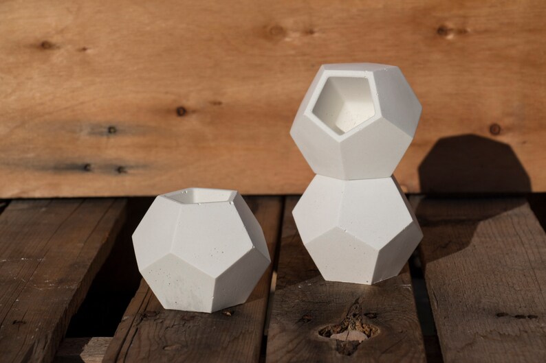 May include: Three white geometric concrete planters of different sizes. The planters are stacked on top of each other. The planters are sitting on a wooden surface.