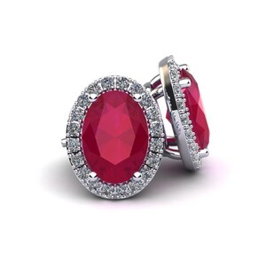 Op de afbeelding: Een paar robijnrode oorstekers. Elke oorbel heeft een ovaal geslepen, levendige rode robijn, omringd door een halo van kleine diamanten. De oorbellen zijn gezet in een zilverkleurig metaal, wat een klassieke en elegante uitstraling geeft.