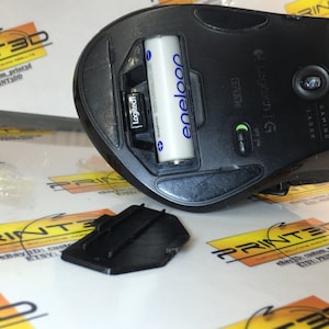 May include: A black Logitech G gaming mouse with a battery compartment open, revealing a blue eneloop battery. The mouse has a green on/off switch and the text "Logitech G" and "Gaming Lager" printed on the side.