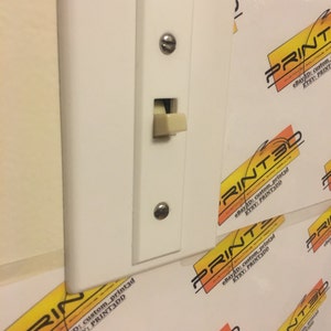 Könnte beinhalten: Ein weißer Lichtschalter mit einem beigefarbenen Kippschalter ist an einer Wand montiert. Der Schalter ist rechteckig mit einem kleinen, rechteckigen Kippschalter. Die Wand ist mit einem sich wiederholenden Muster aus Logos in Orange, Schwarz und Weiß bedeckt.
