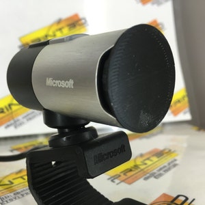 May include: A silver and black Microsoft webcam with a clip for attaching to a monitor. The webcam has a black lens cover.