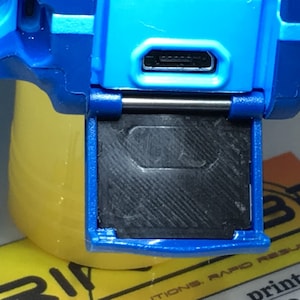 May include: Close-up of a blue device with a black flap covering a slot. The flap is open, revealing a black slot. A USB-C port is visible on the device.
