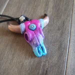 May include: A handcrafted bull skull pendant with a gradient of pink, purple, and turquoise hues. The skull features a textured eye detail and is strung on a black cord. The horns are a light brown color.