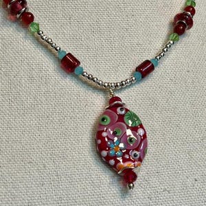 May include: A red, white, and green glass pendant with a floral design hangs from a silver chain necklace with red and blue beads.