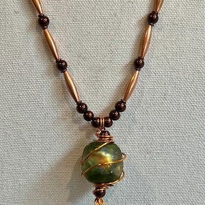 Puede incluir: Collar hecho a mano con una cuenta de vidrio verde y dorado envuelta en alambre de cobre. El collar tiene cuentas tubulares de color cobre y pequeñas cuentas marrones. El collar se muestra sobre un fondo azul claro.