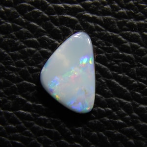 May include: A polished, pear-shaped opal gemstone. The opal has a milky white base color with iridescent flashes of blue, green, and pink. The stone is set against a black, textured background.
