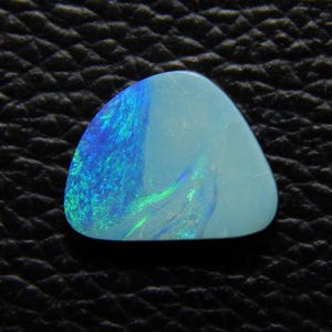 May include: A polished, triangular opal gemstone. The opal displays a mix of light blue and vibrant blue-green colors, with a translucent quality. The stone is set against a dark, textured background.