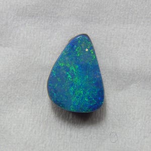May include: A polished, teardrop-shaped opal with a vibrant display of color. The gemstone features a deep blue base with flashes of green and turquoise throughout. The opal is set against a neutral background.