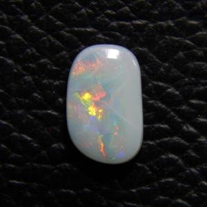 May include: A polished, oval-shaped opal gemstone. The opal is primarily white with iridescent flashes of yellow, orange, and green. The gemstone is set against a dark, textured background.