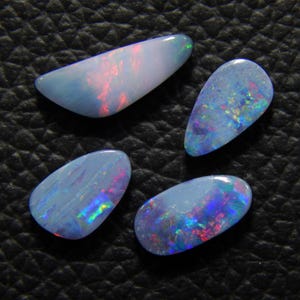 May include: Four polished opal gemstones with a blue base color and iridescent flashes of red, green, and purple. The opals have a smooth, glossy surface and are set against a dark, textured background. Each stone has a unique shape.