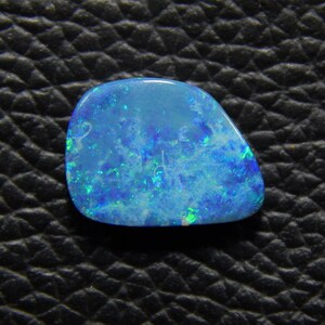 May include: A polished, oval-shaped opal gemstone. The opal displays a vibrant play-of-color with shades of blue and green, set against a translucent background. The gemstone is displayed on a textured black surface.