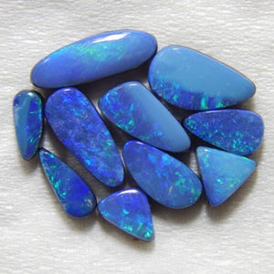 May include: A collection of polished opals in various shapes, showcasing a range of blue hues with flashes of green. The opals are smooth and glossy, with a play of color visible on their surfaces. The gemstones are arranged on a white background.