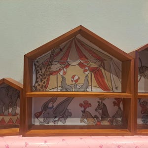 May include: A wooden shelf unit with three house-shaped compartments. Each section displays a circus scene, such as an elephant on a stand, seals with balls, and a tiger on a tightrope. The unit is crafted from warm-toned wood.