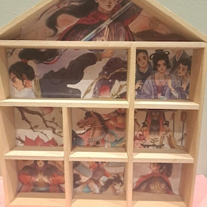 May include: Wooden shelf unit with nine compartments, each displaying a different illustration of Asian-inspired figures. The illustrations feature people in traditional clothing, some on horseback, in a variety of poses and settings. The wood is unfinished.