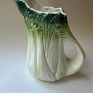 Może przedstawiać: Biały ceramiczny dzbanek w kształcie selera z zielonymi liśćmi. Dzbanek ma uchwyt i jest przeznaczony do przechowywania płynów.