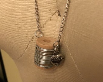 Puede incluir: Un collar de cadena plateada con un dije de carrete de madera y un dije de corazón plateado que dice "Made with Love".