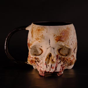 Pode incluir: Uma caneca de cerâmica branca em forma de crânio com uma alça preta. O crânio tem respingos de tinta vermelha e marrom, o que lhe confere uma aparência sangrenta.